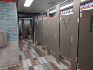 Brown bathroom stalls with brown roman numerals and metal join accents atop of a multi-colored rectangular floor tiles.